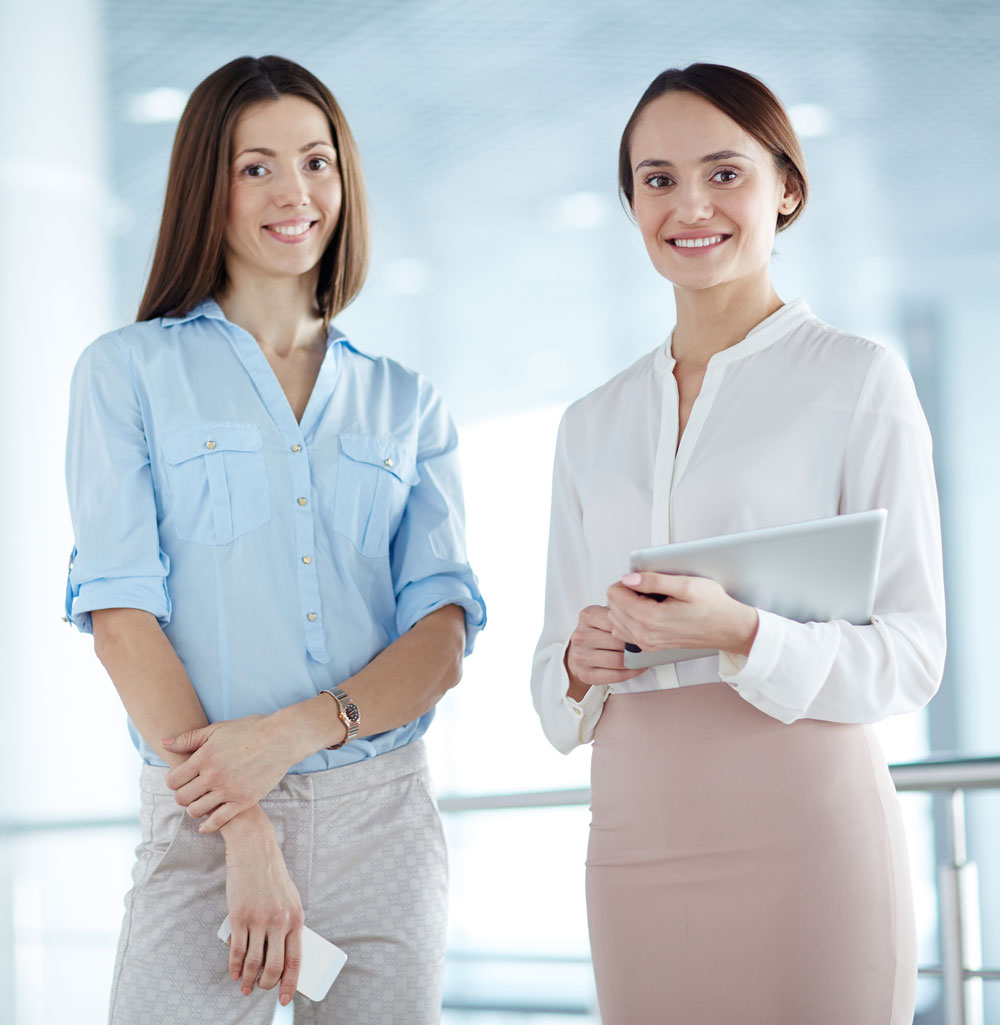 Two professional women smiling together in a bright, modern office, representing connection, collaboration, and women in business.
